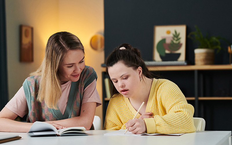 Young Caucasian woman spending time with her younger sister with Down syndrome helping with homework task